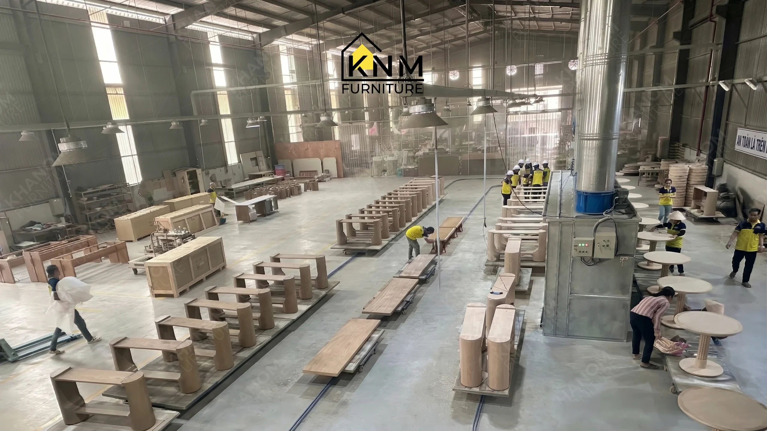 Dozens of tables on a continuous trolley track moving towards a large paint booth and packing area at Khanom Furniture (KNM Furniture), a hospitality furniture factory Vietnam.