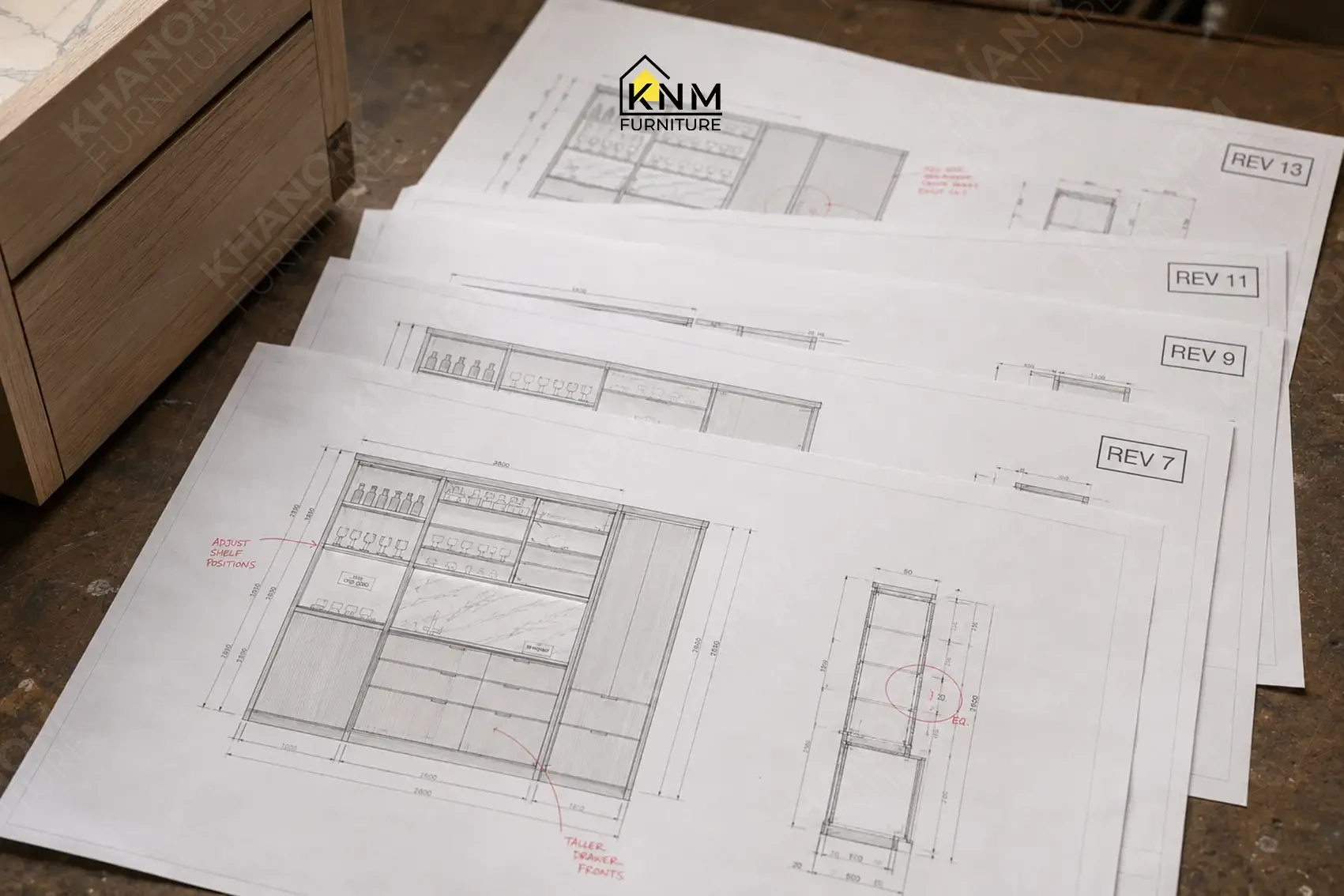 Multiple printed technical drawing revisions spread on a table alongside a wooden cabinet sample at Khanom Furniture (KNM Furniture), a custom furniture factory Vietnam.