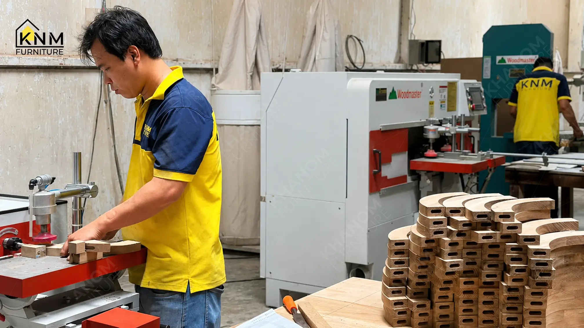 Khanom Furniture craftsman operating CNC woodworking machinery for chair frame component batch production, stacked solid hardwood parts visible in foreground, second craftsman at machine in background.
