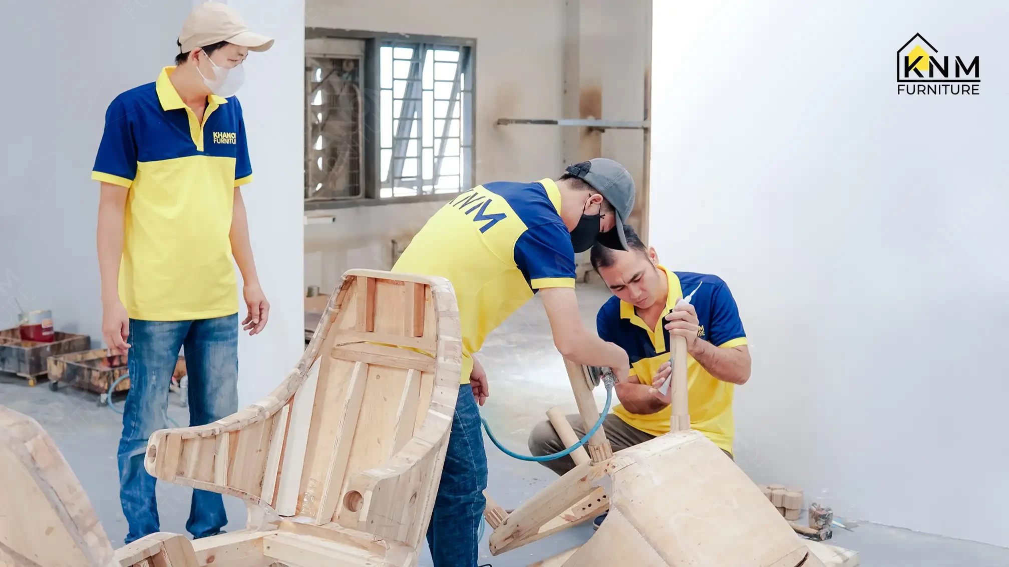 Three Khanom Furniture (KNM Furniture) craftsmen in yellow-navy uniforms assembling a solid hardwood armchair frame using a pneumatic staple gun and hand tools in a production workshop
