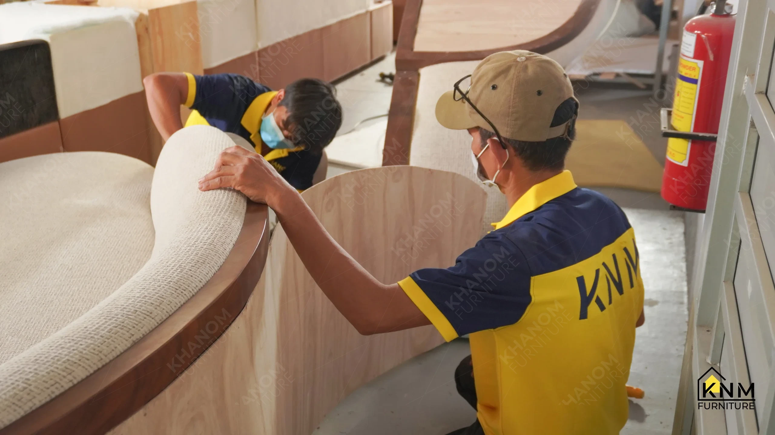 Khanom Furniture (KNM Furniture) craftsmen applying cream fabric upholstery to a large curved wood-framed sofa during assembly, factory workshop Vietnam