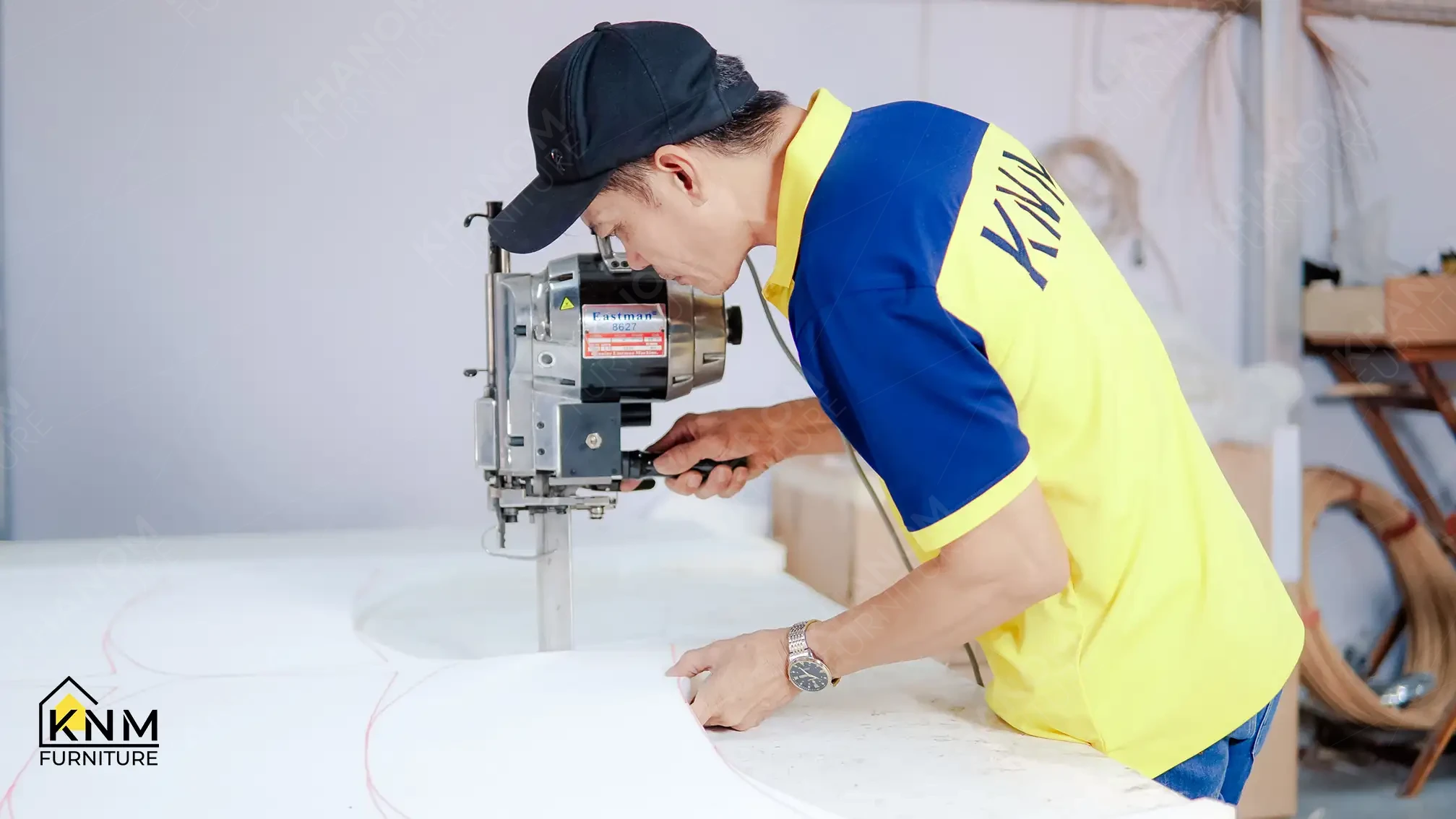 KNM upholstery craftsman operating an Eastman Model 8627 straight-knife cutting machine to cut white batting or foam sheeting on a large cutting table.