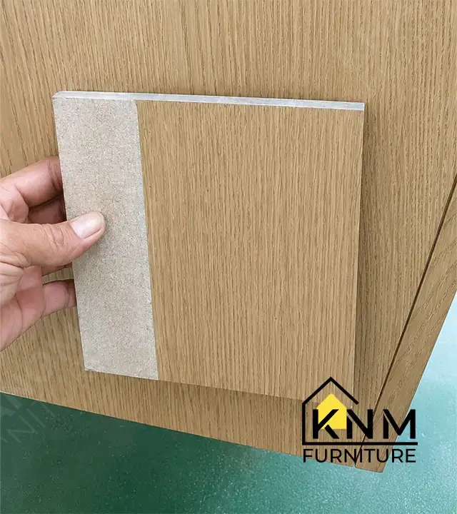 Khanom Furniture quality inspector holding an approved veneer finish swatch sample directly against a completed casegood door panel for color and grain comparison during pre-shipment QC.