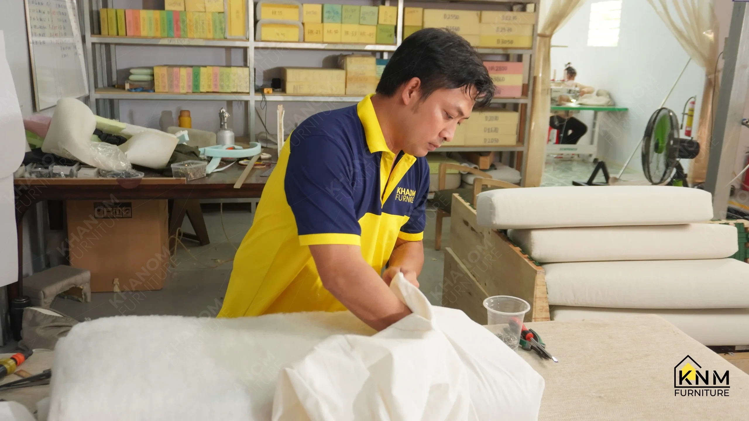 Khanom Furniture (KNM Furniture) craftsman fitting fabric over a foam cushion insert, graded foam density blocks labeled by code visible on factory shelving in background