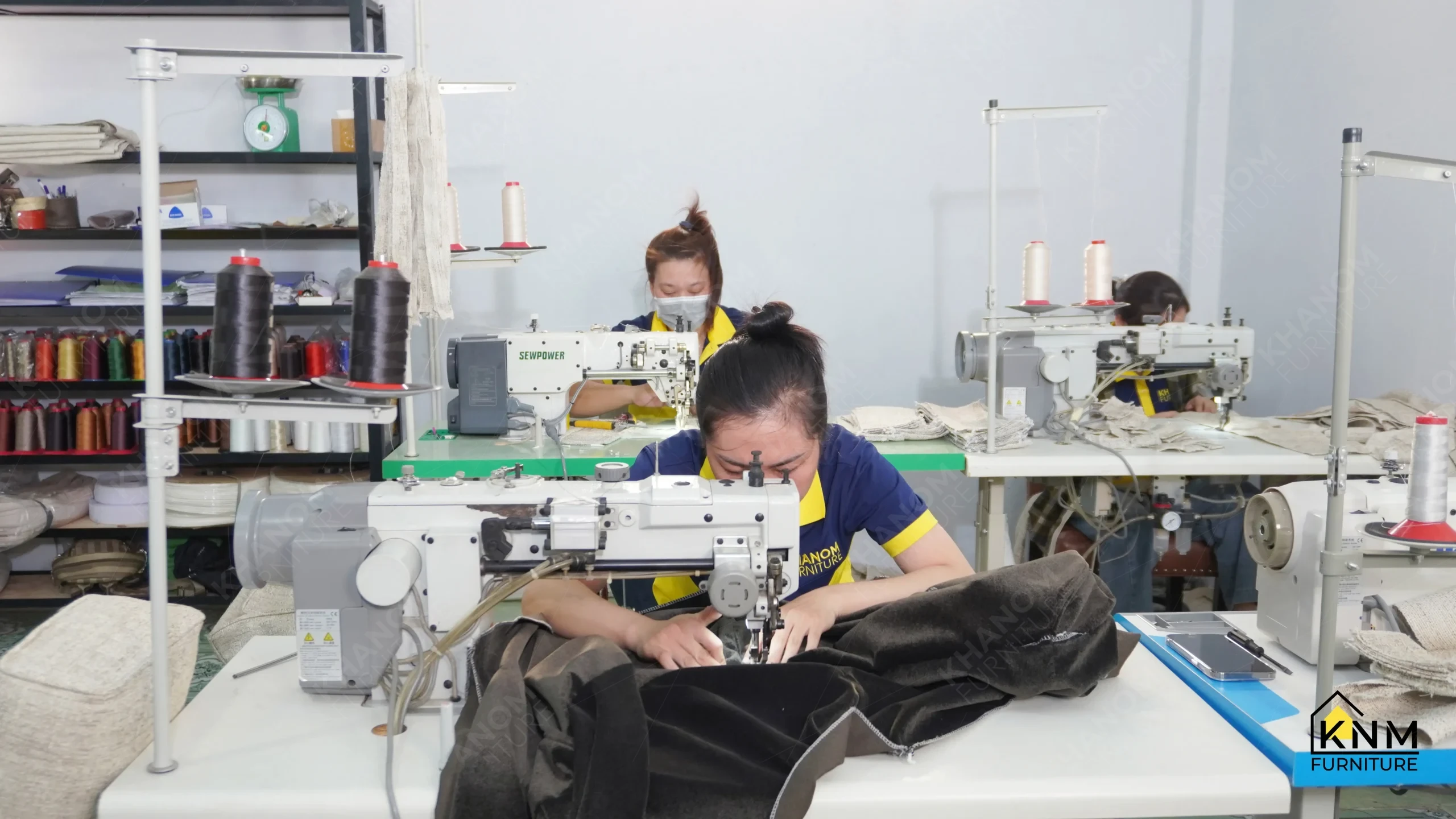 Khanom Furniture (KNM Furniture) upholstery staff operating industrial single-needle sewing machines to construct fabric panels, thread spools and material bolts visible on shelving