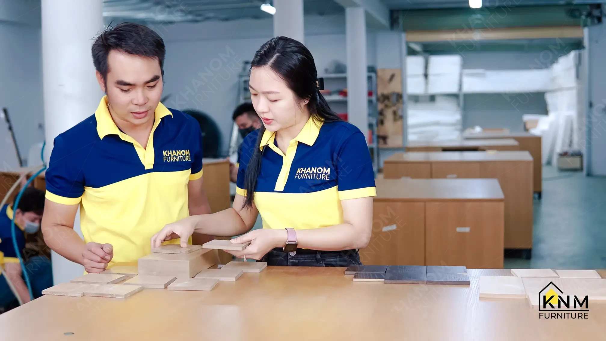 Two Khanom Furniture staff in yellow-navy uniforms reviewing multiple wood veneer finish samples on a table, with completed casegoods visible in background staging area.