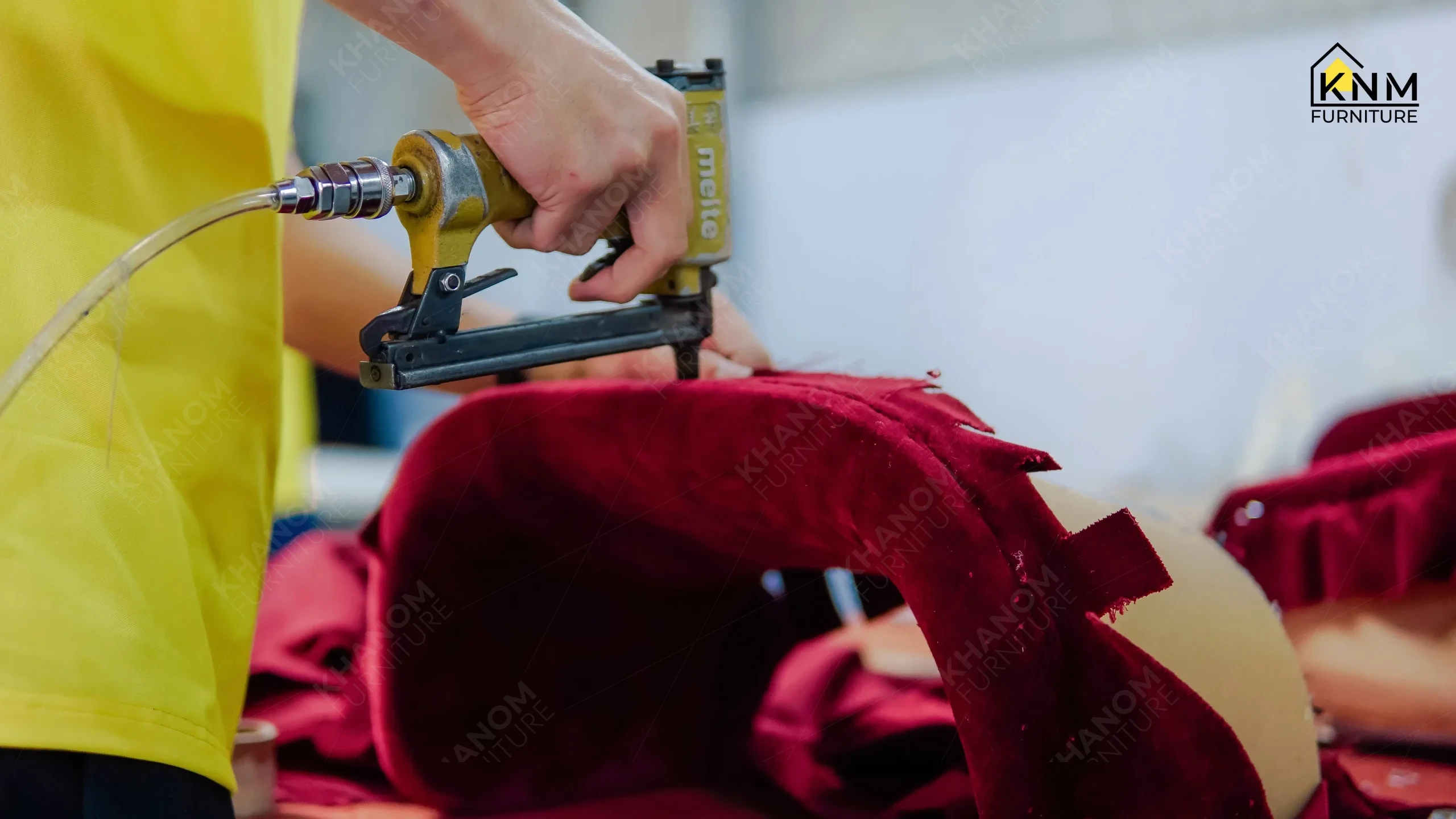 Close-up of a Khanom Furniture (KNM Furniture) craftsman using a pneumatic staple gun to tension maroon velvet fabric onto a chair frame edge during upholstery application