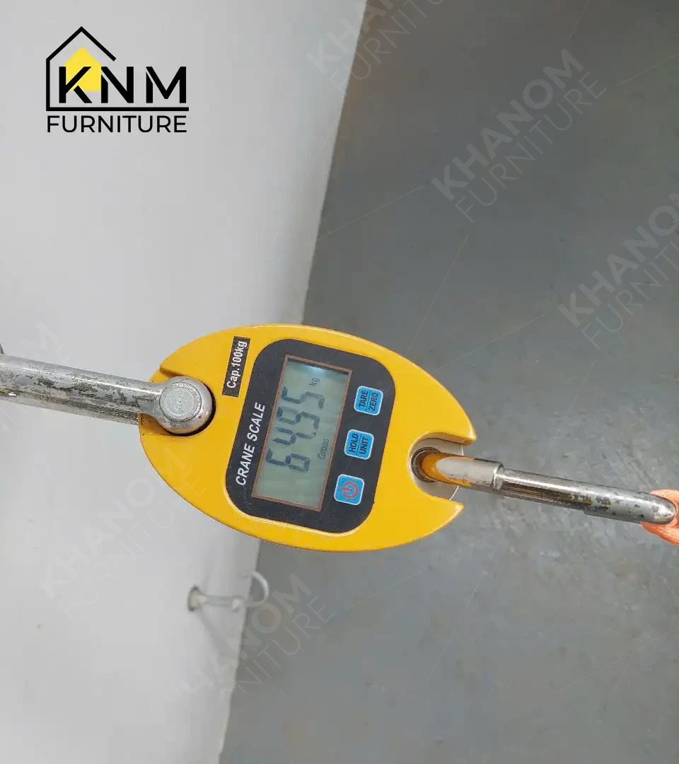 Yellow digital crane scale (100 kg capacity) recording applied load during furniture structural testing at KNM Furniture, KNM logo visible in upper corner.