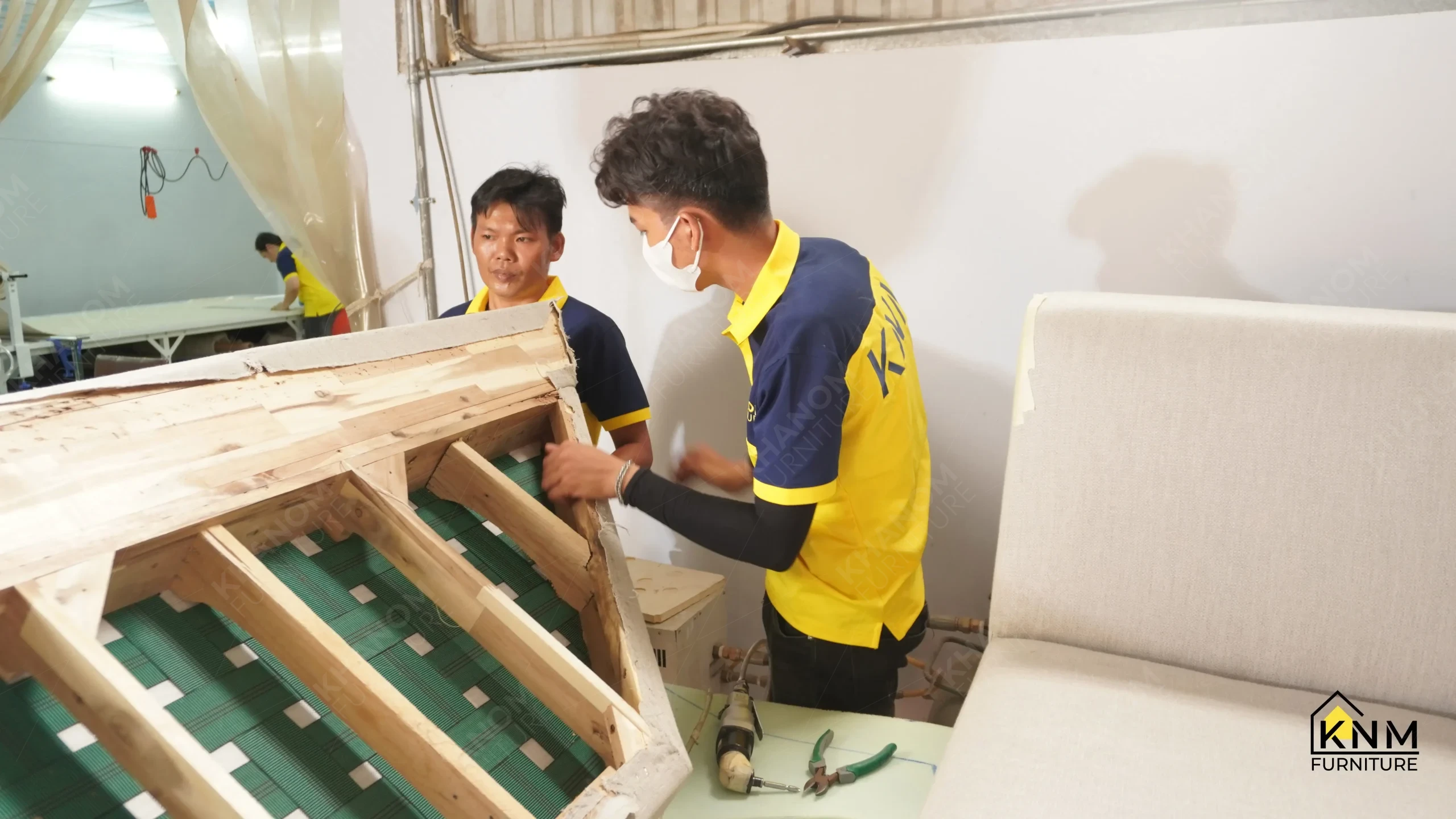 Khanom Furniture (KNM Furniture) craftsmen inspecting the webbing suspension system on an overturned sofa frame before foam application, upholstery factory Vietnam