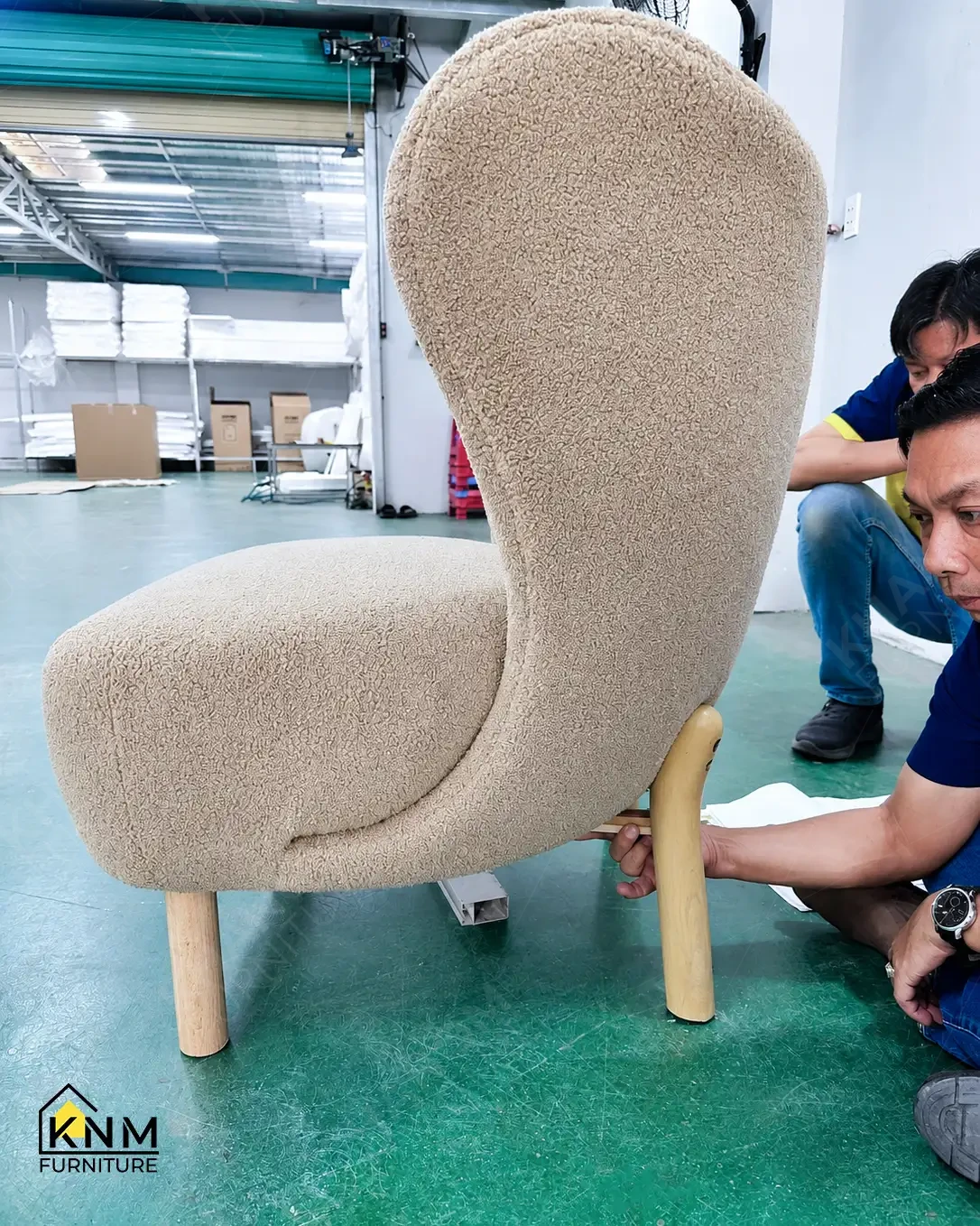 Khanom Furniture (KNM Furniture) craftsmen attaching turned wood legs to a finished boucle-upholstered lounge chair as a final assembly step, factory floor Vietnam